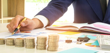 Businessman has a pen in his right hand analysis of graph data,On the desk there are coins, dollars, notebooks and flip charts placed.